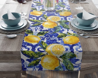 May include: A white table runner with a blue and white tile pattern featuring lemons and green leaves. The runner is on a wooden table with place settings.
