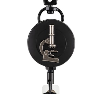 以下が含まれることがあります： 銀色の顕微鏡のデザインが施された黒いリトラクタブルバッジリール。リールには黒いクリップと白いプラスチック製のバッジホルダーが付いています。