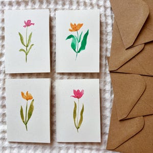 May include: Four watercolor greeting cards with floral designs in pink and orange, accompanied by brown paper envelopes. The cards feature hand-painted flowers with green stems and leaves on a white background.