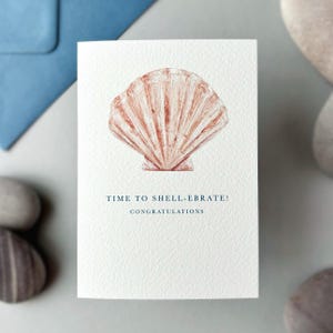 May include: A white greeting card featuring a peach and brown seashell illustration. Navy blue text reads "TIME TO SHELL-EBRATE! CONGRATULATIONS". A blue envelope is visible in the background. The card is a celebration-themed design.