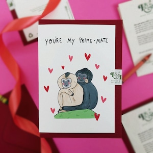 May include: A white card with a red border and the text "You're my prime-mate" in black. The card features a cartoon illustration of two gibbons hugging, surrounded by red hearts.