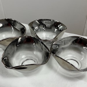 May include: Four silver-rimmed glass bowls with a dark gray interior. The bowls are arranged in a semi-circle on a white surface.