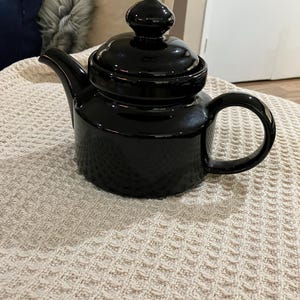 May include: A black ceramic teapot with a shiny finish and a rounded body. The teapot has a lid with a knob on top and a spout on one side. The handle is on the opposite side of the spout.