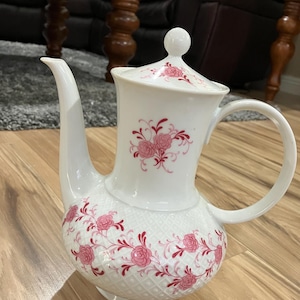 May include: White porcelain teapot with a pink and red floral design. The teapot has a lid and a handle.