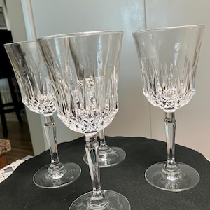 Może przedstawiać: Cztery przezroczyste kryształowe kieliszki do wina ze skomplikowanymi wzorami ciętego szkła. Kieliszki mają klasyczny kształt z okrągłą podstawą, smukłą nóżką i czaszą z pionowymi i rombowymi cięciami. Ustawione na ciemnej powierzchni.