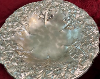 May include: A silver-colored, round decorative serving dish with a floral and leaf design around the rim. The dish has a shallow, concave center and is likely made of metal.