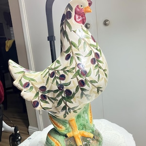 May include: A ceramic hen figurine with a cream-colored body, adorned with olive branch patterns. The hen has a red comb and wattle, an orange beak, and stands on a green base. The figurine is a decorative item.