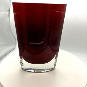 May include: A deep red glass vase with a thick, clear glass base. The vase features a smooth, slightly tapered cylindrical shape with vertical interior indentations that create a subtle, rhythmic pattern within the translucent red glass walls.
