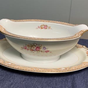 May include: A white porcelain gravy boat with a matching oval-shaped underplate. The set features a floral design with pink, yellow, and purple flowers, and gold trim. The gravy boat has a handle on each side.