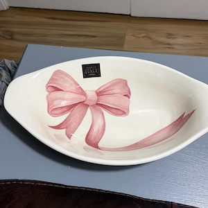 May include: An oval-shaped white ceramic serving dish featuring a hand-painted pink ribbon bow design in the center. A small black rectangular label with gold text reading ITALY is positioned near the top rim of the interior of the bowl.