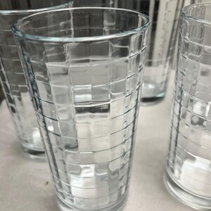 May include: A set of clear glass tumblers featuring a raised grid pattern across the exterior surface. Each cylindrical drinking glass has a smooth rim and a thick, flat base, creating a textured, geometric design that catches the light.