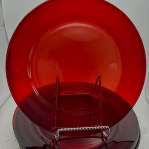 May include: A stack of circular, translucent red glass plates. The top plate is positioned upright on a silver wire display stand, showcasing a smooth, glossy surface and a simple, rimmed edge design. The plates are uniform in color and shape.