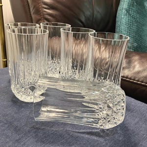 May include: A set of six clear, cut-glass tumblers. The glasses have vertical lines on the sides and diamond-shaped patterns near the base. One glass is lying on its side. The glasses are on a blue surface.