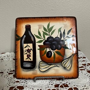 May include: A square ceramic serving plate with a painted design of a bottle of olive oil, a bowl of olives, and garlic cloves. The plate has a brown border and a cream background.