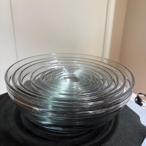 May include: A set of clear glass bowls nested inside one another, featuring a smooth, rounded design with a transparent finish. The stack consists of multiple circular bowls of varying sizes, creating a layered, concentric appearance.