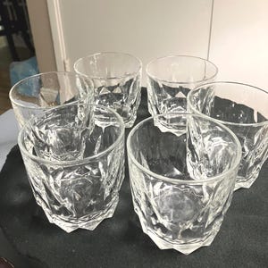 May include: A set of six clear glass tumblers featuring a geometric, faceted diamond-like pattern on the exterior. Each glass has a thick base and a wide, circular rim, designed with a heavy-bottomed structure for stability and a textured surface.
