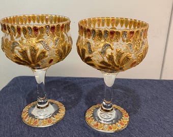 May include: Two ornate, clear glass goblets with gold, red, and silver accents. The bowls have a textured, floral design, and the rims and bases are decorated with a repeating pattern. The stems are clear, and the goblets sit on a blue surface.
