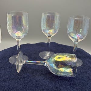 May include: A set of four clear glass wine glasses with an iridescent finish that reflects rainbow colors. Three glasses stand upright on thin stems with circular bases, while one glass lies horizontally in the foreground on its side.