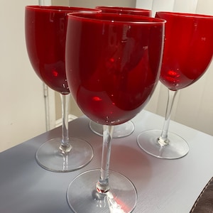 May include: Four red wine glasses with clear stems and bases. The glasses have a rounded bowl shape and are made of a glossy, transparent material. The glasses are arranged on a light gray surface.