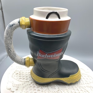 May include: A novelty ceramic mug shaped like a firefighter's boot, with a hose-like handle. The mug is black, with a yellow sole and accents, and features the Budweiser logo in red and white.
