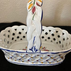 May include: White ceramic basket with a woven design and a braided handle. The handle is decorated with painted flowers in red, yellow, and green. The basket has a blue and white trim.