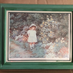 Puede incluir: Una impresión enmarcada de una niña pequeña con un vestido blanco y un sombrero de paja de pie en un jardín con flores amarillas. La niña sostiene un aro en su mano.
