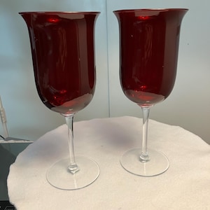 May include: Two tall, ruby red wine glasses with a tulip-shaped bowl and a slender stem. The glasses are made of transparent glass and sit on a clear, circular base. The glasses are displayed on a white surface.