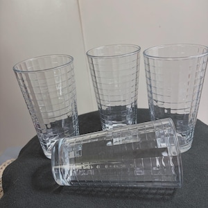 May include: A set of four clear glass tumblers, each featuring a textured grid pattern of raised squares across the exterior surface. Three of the glasses are standing upright, while one glass is positioned horizontally in the foreground.