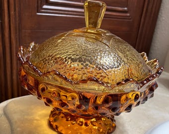 May include: A vintage amber glass candy dish with a domed lid. The dish has a scalloped edge and a textured, bubbled surface. The base of the dish is decorated with small, raised dots.