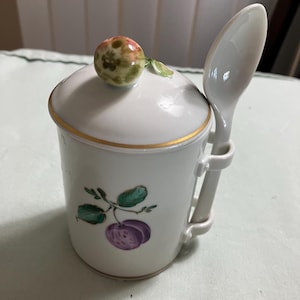 May include: White porcelain sugar bowl with a lid and a spoon. The lid has a floral design and a small fruit on top. The bowl has a floral design and a purple fruit on the side. The spoon is attached to the side of the bowl.