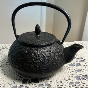 Puede incluir: Tetera de hierro fundido negro con una superficie texturizada y un asa curva. La tetera tiene una tapa con un pequeño pomo y un pico para verter. La tetera está sobre un tapete de ganchillo blanco.