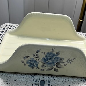May include: Cream-colored ceramic napkin holder with a rectangular base and curved sides. The front features a floral design with blue roses and gray leaves. The holder has a raised back for support.