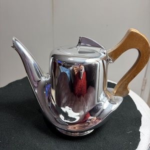 May include: A shiny silver teapot with a wooden handle. The teapot features a curved spout and a lid with a small fin. The handle is a light brown colour. The teapot is on a black surface.