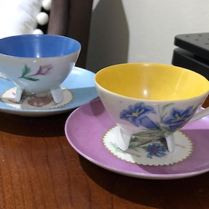 May include: Two teacups with floral designs and matching saucers. The teacups have three legs and are decorated with blue and yellow flowers. The saucers are blue and purple.