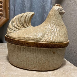 May include: A ceramic chicken-shaped casserole dish with a brown and beige glaze. The dish has a ribbed design and a lid shaped like a hen.