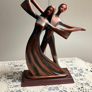 Puede incluir: Escultura de bronce de una pareja bailando. Las figuras están estilizadas con líneas fluidas, brazos extendidos y una sensación de movimiento. La escultura está montada sobre una base rectangular.