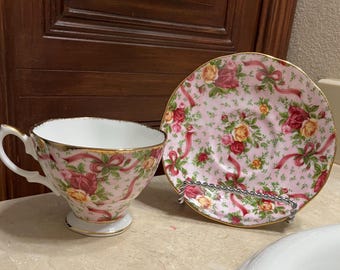 Könnte beinhalten: Ein rosa und goldenes Tee-Set mit Blumenmuster. Tasse und Untertasse sind mit rosa Rosen, grünen Blättern und goldenen Akzenten verziert.