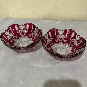May include: Two small, clear glass bowls with a deep burgundy red tint. The bowls have a scalloped edge and are decorated with a white, etched design of hearts and flowers.