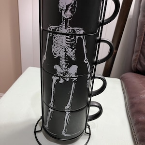May include: Set of four black ceramic mugs with a skeleton design. The mugs are stacked on a metal stand.