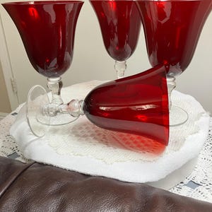 May include: Four red wine glasses with clear stems and bases. One glass is lying on its side, while the other three stand upright. The glasses have a tulip-shaped bowl and are set on a white, lacy surface.