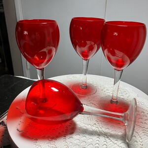 May include: Four red wine glasses with clear stems and bases. One glass is lying on its side, while the other three stand upright on a white, lace-covered surface. The glasses have a rounded bowl shape.
