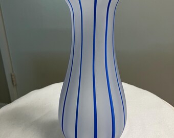 May include: A frosted glass vase with vertical blue stripes. The vase has a flared top and a rounded base. The blue stripes are evenly spaced and run the length of the vase, creating a decorative pattern. The vase is sitting on a white surface.
