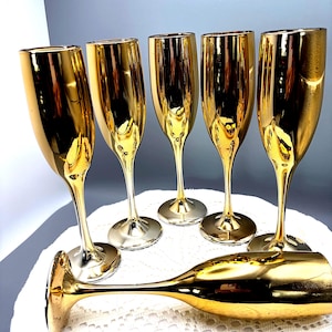 May include: A set of six champagne flutes with a reflective gold metallic finish. Five of the flutes are standing upright in a semi-circular arrangement, while one flute is positioned horizontally in the foreground, showcasing the tall stem.