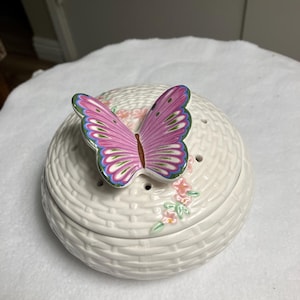 May include: A white ceramic trinket box with a woven basket design. The lid is adorned with a pink, purple, and green butterfly figurine. Delicate pink floral accents are painted on the side of the box.