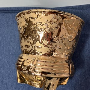 May include: A gold ceramic planter with a textured, crackled glaze. The planter has a wide, round opening and a base with three legs.