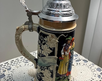 May include: A ceramic beer stein with a hinged, silver-coloured lid. The stein features a relief design of figures in traditional clothing, with a handle and decorative relief. The background is dark blue with cream-coloured relief.
