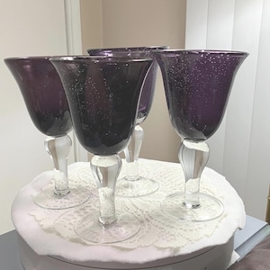 May include: Four elegant wine glasses with a deep purple hue. The glasses have a flared bowl design with a clear, decorative stem and base. The purple glass has a textured, bubbly appearance. The glasses are arranged on a white, lacy surface.