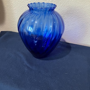 May include: A cobalt blue glass vase with a ruffled rim and a spiral design. The vase has a rounded shape and is made of transparent glass. The vase is sitting on a dark blue surface.