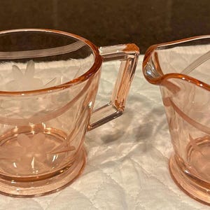 May include: A pink glass sugar bowl and creamer set. The sugar bowl has two handles and the creamer has a spout. Both pieces have etched floral and line designs. The set is made of transparent glass.