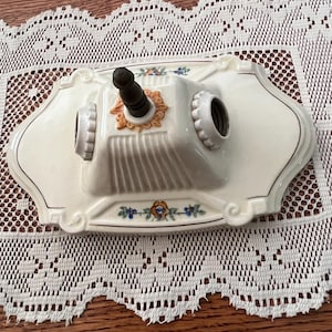 May include: Cream-colored ceramic wall sconce with a decorative floral design. The sconce features two light sockets and a central candle holder. The edges are outlined with a brown trim. The sconce is placed on a white lace doily.